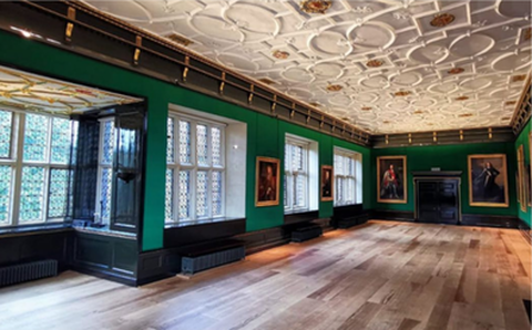 A grand hall from Tudor times. The photo is taken from one corner looking down the length of the room. On the left are large panelled windows divided up into small diaganol grids. They are surrounded by black wood and ornate white ceilings. The rest of the room is painted a forest green. On each part of the wall is a large portrait of dignatories of old, surrounded in ornate gilt frames. The whole of the ceiling is covered in an ornate carved white grid of circles, foliage and an occasional gold leafed flower.