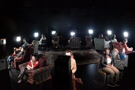 Fourteen young adults sitting in a circle with their backs to each other. In the middle of them is a large patterned rug. Each person sits on a different kind of chair, some hard, some soft. In front of each person is a tv screen, and everyone is wearing headphones. The lighting is very dark except for the light coming from the screens.