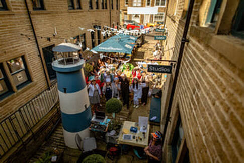 The camera angle is high up, looking down on a celebratory and creative scene. Nestled between two tall sandstone terraces is a tall blue and white striped lighthouse. Behind it a crowd of people stands under sunshades and bunting.